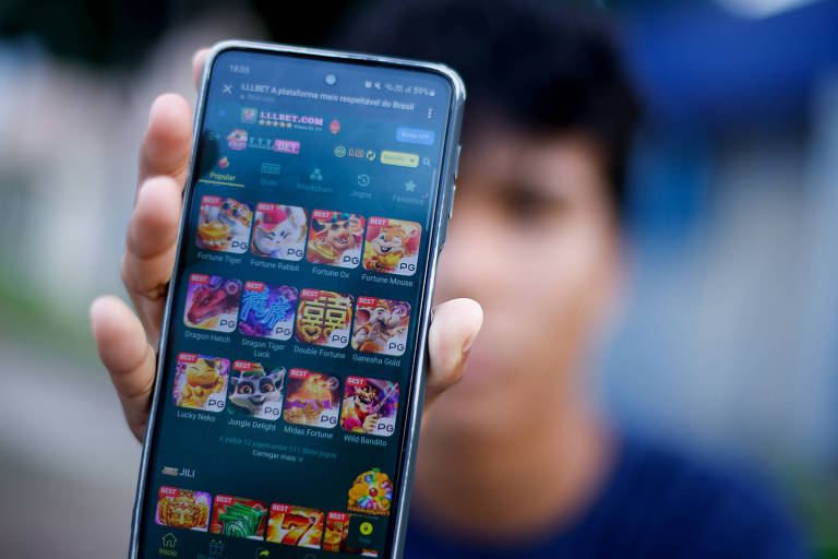 A imagem mostra uma pessoa segurando um smartphone com a tela voltada para a câmera. Na tela do smartphone, há vários ícones de jogos coloridos organizados em uma grade. A pessoa está desfocada ao fundo, enquanto o smartphone está em foco.