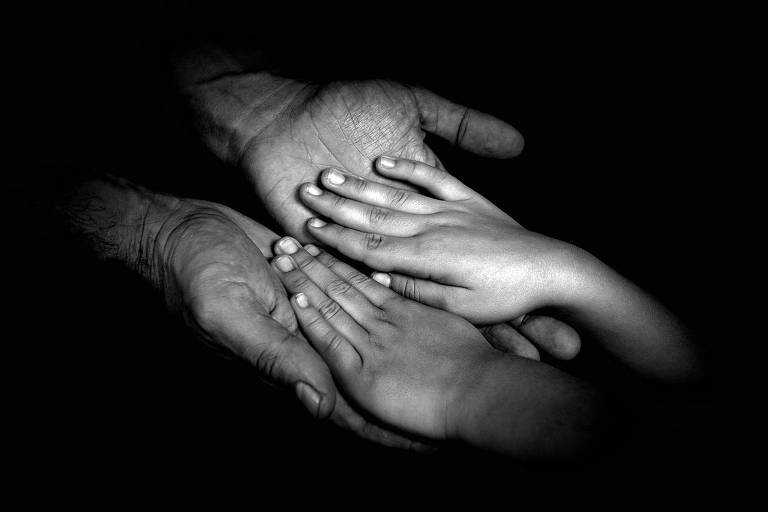 A imagem em preto e branco mostra duas mãos adultas segurando duas mãos infantis, simbolizando proteção e cuidado. As mãos estão em destaque contra um fundo preto, enfatizando o contraste e a conexão entre as gerações.