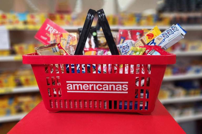 Uma cesta de compras vermelha da rede 'Americanas' está cheia de produtos, como alimentos e bebidas. A cesta está posicionada em uma superfície vermelha, com prateleiras de produtos ao fundo, desfocadas.