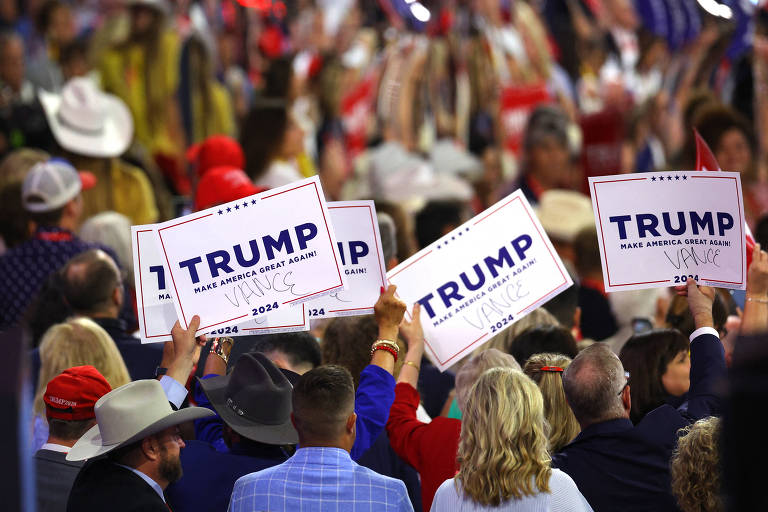A imagem mostra um grupo de pessoas em um evento, segurando placas que dizem 'TRUMP' em letras maiúsculas. As placas são brancas com texto azul. A multidão está vestida de forma variada, com algumas pessoas usando chapéus.