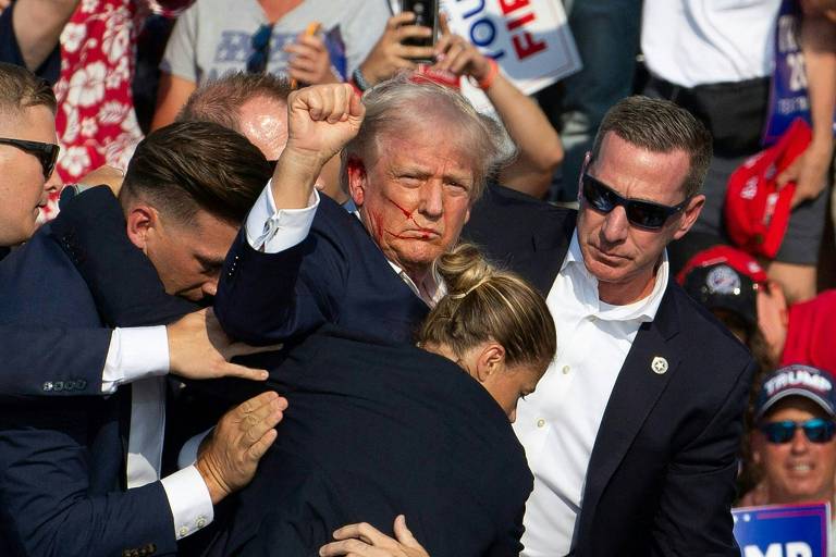 A imagem mostra Trump, de terno azul, levantando o punho em meio a uma multidão. O rosto dele está sujo de sangue. Ele está cercado por várias pessoas, incluindo seguranças que tentam protege-lo. Ao fundo, há várias pessoas vestindo roupas casuais, algumas segurando cartazes e bandeiras.