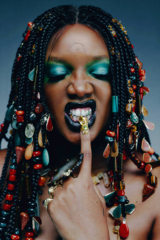 A imagem mostra a cantora Iza com maquiagem vibrante, incluindo sombra de olhos verde brilhante e batom preto. Ela está usando tranças decoradas com contas coloridas e acessórios. E está com um dedo na boca, exibindo um anel dourado em forma de garra.