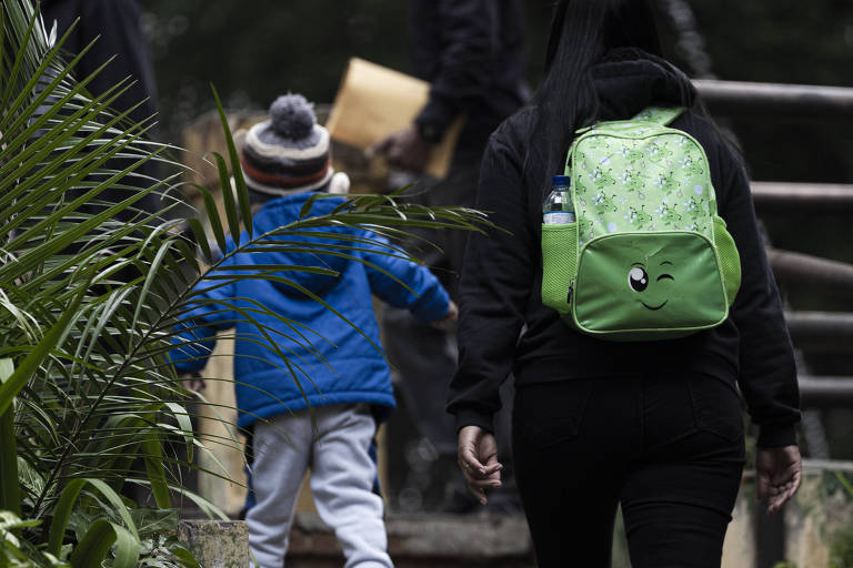 A imagem mostra uma criança vestindo um casaco azul e um gorro cinza, caminhando ao lado de um adulto que usa um moletom preto e carrega uma mochila verde. Eles estão ao ar livre, cercados por vegetação, e parecem estar subindo uma escada.