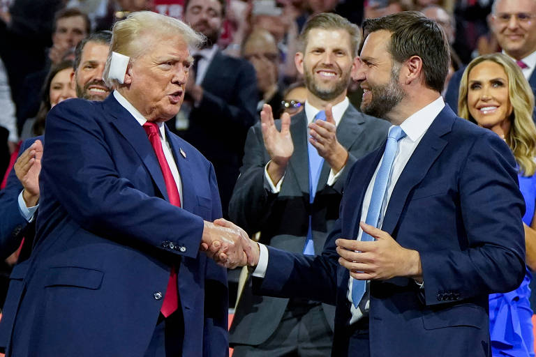 Dois homens estão apertando as mãos em um evento. O homem à esquerda está vestindo um terno azul escuro com uma gravata vermelha, enquanto o homem à direita está vestindo um terno azul com uma gravata azul clara. Ao fundo, várias pessoas estão aplaudindo e sorrindo.