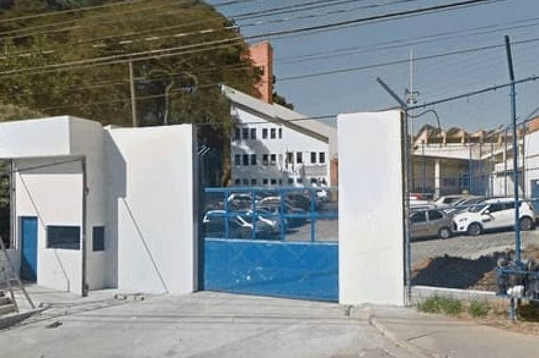 A imagem mostra a entrada de um estacionamento cercado por um portão azul e muros brancos. Há vários carros estacionados no interior, e ao fundo, é possível ver edifícios brancos com janelas