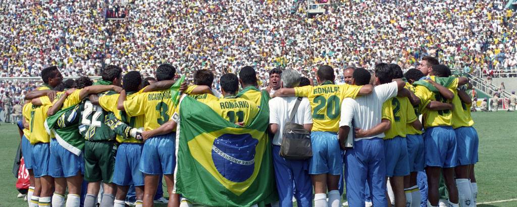 Imagem de uma equipe de futebol brasileira em círculo, abraçados, no campo. Os jogadores estão vestindo uniformes amarelos e azuis, com alguns segurando uma bandeira do Brasil. Ao fundo, uma grande multidão de torcedores está presente nas arquibancadas.