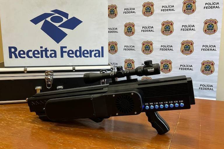 A imagem mostra uma arma de fogo preta com uma mira telescópica, colocada sobre uma mesa de madeira. Ao fundo, há um banner com os logotipos da Polícia Federal e da Receita Federal, além de uma maleta preta com o logotipo da Receita Federal.
