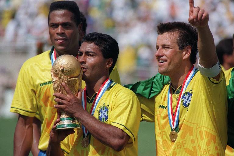 Três jogadores de futebol vestindo uniformes amarelos da seleção brasileira estão comemorando com a taça da Copa do Mundo. O jogador no centro está segurando a taça e beijando-a, enquanto os outros dois jogadores estão ao seu lado, um deles apontando para o céu. Todos estão usando medalhas no pescoço.