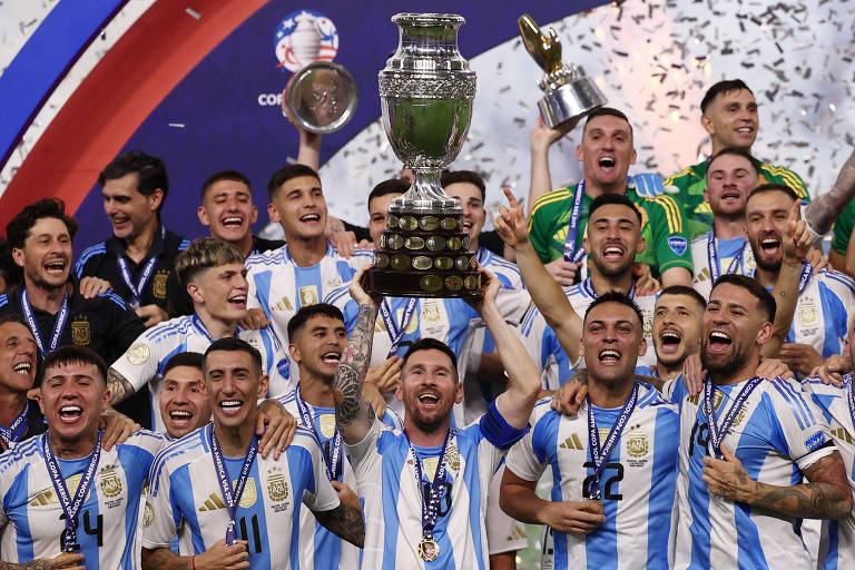 A imagem mostra a equipe de futebol argentina comemorando uma vitória. Os jogadores estão vestindo uniformes listrados em azul e branco, com medalhas penduradas no pescoço. Um jogador no centro está levantando um grande troféu prateado, enquanto outros jogadores ao redor sorriem e levantam os braços em celebração. Ao fundo, há confetes caindo e um banner com o logotipo da Copa América.