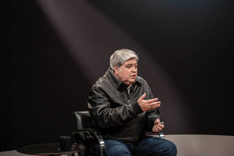 Um homem de cabelos grisalhos, usando uma jaqueta preta, está sentado em uma cadeira de rodas. Ele gesticula com as mãos enquanto fala, em um ambiente de estúdio com fundo escuro e uma luz suave incidindo sobre ele.