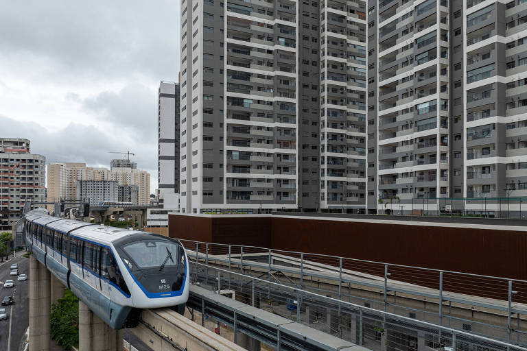 A imagem mostra um trem moderno de cor azul e branca passando por uma linha elevada em uma área urbana. Ao fundo, há vários edifícios residenciais altos com muitas janelas e varandas. O céu está nublado.