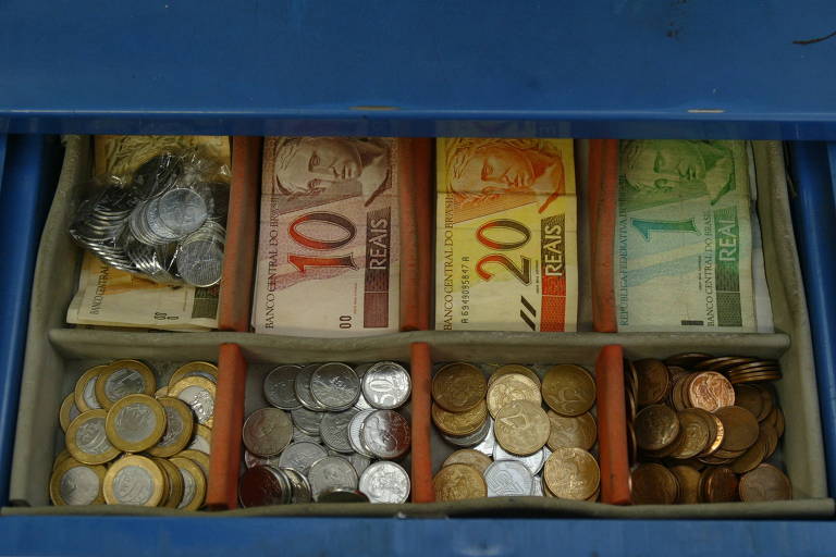 Imagem de uma gaveta de caixa registradora contendo cédulas e moedas de real brasileiro. As cédulas estão organizadas em compartimentos separados e incluem notas de 1, 10 e 20 reais
