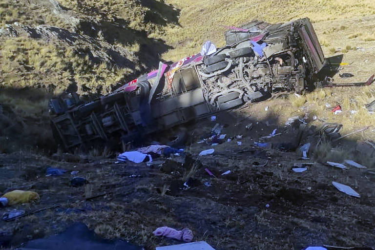 A imagem mostra um ônibus tombado de lado em uma área montanhosa. O veículo está parcialmente destruído e há destroços espalhados ao redor, incluindo roupas e outros objetos pessoais. A área ao redor é composta por vegetação rasteira e terreno acidentado.