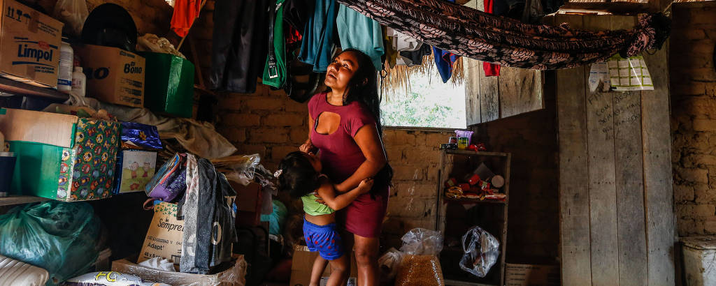 A imagem mostra o interior de uma casa simples com paredes de tijolos e teto de palha. Uma mulher está de pé, sorrindo, enquanto segura uma criança. Ao redor, há várias caixas, sacolas e roupas penduradas.