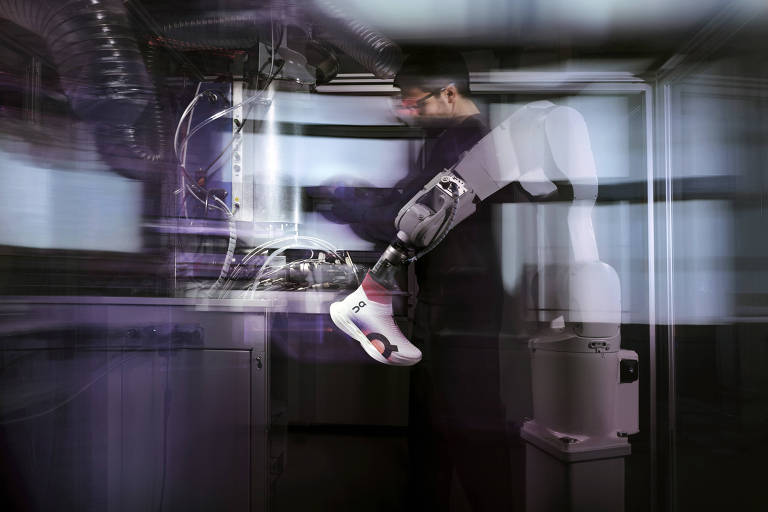 A imagem mostra um robô industrial manipulando um tênis em um ambiente de laboratório. O robô possui um braço mecânico articulado que segura o tênis, enquanto um homem ao fundo parece estar operando ou monitorando o processo. O ambiente é preenchido com equipamentos e cabos, sugerindo um cenário de alta tecnologia e automação.