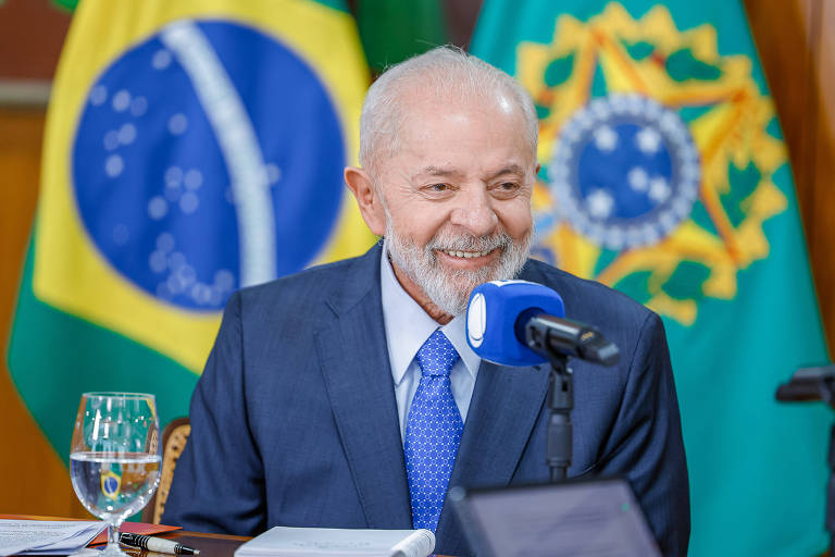 Lula, um homem idoso, de barba branca e cabelo curto, está sorrindo enquanto fala ao microfone. Ele veste um terno azul e uma gravata azul clara. Atrás dele, há uma bandeira do Brasil e outra do brasão da República. Na mesa à sua frente, há um copo de água e alguns papéis.