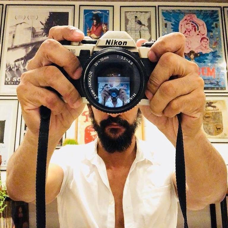 Imagem de um homem com barba segurando uma câmera Nikon em frente ao rosto