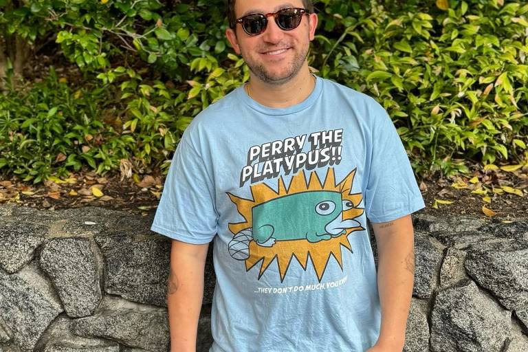 Homem usando óculos de sol e uma camiseta azul clara com a imagem de Perry o Ornitorrinco, personagem de desenho animado. A camiseta tem o texto 'Perry the Platypus!!' e '...They don't do much you'. Ele está em pé em frente a um muro de pedra com vegetação verde ao fundo.