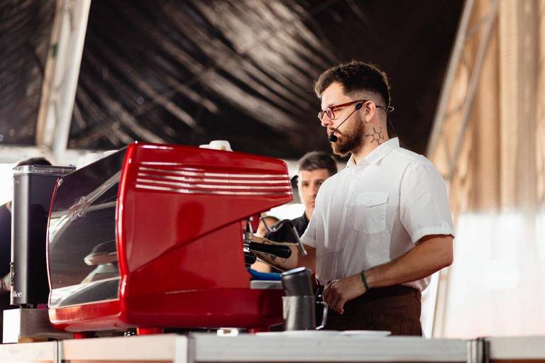 A imagem mostra um barista operando uma máquina de café expresso vermelha. Ele está usando uma camisa branca e um avental marrom, além de um fone de ouvido com microfone. O barista tem uma tatuagem visível no pescoço e usa óculos. Ao fundo, há outra pessoa parcialmente visível e um ambiente com iluminação suave.
