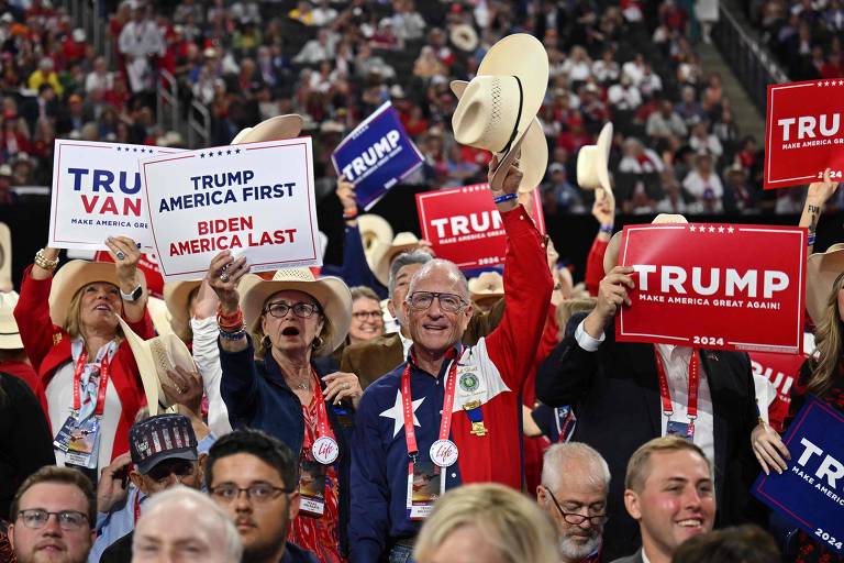 A imagem mostra um grupo de pessoas em um comício político, segurando cartazes de apoio a Donald Trump. Alguns dos cartazes dizem América de Trump em primeiro lugar, América de Biden em último. As pessoas estão vestidas com roupas com as cores nacionais dos Estados Unidos, incluindo chapéus de cowboy e jaquetas vermelhas, e estão sorrindo.