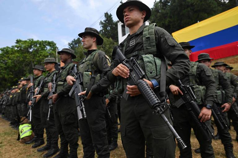 Homens com uniformes verdes seguram armas; há um bandeira da colômbia ao fundo