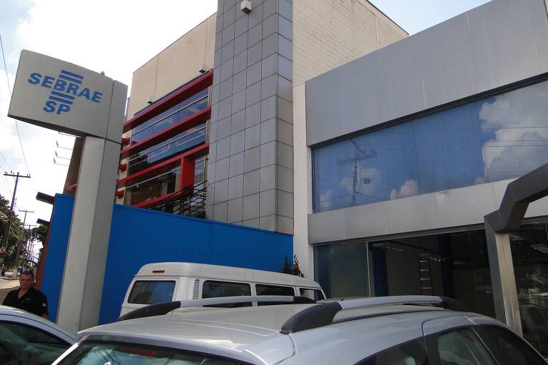 Imagem de uma fachada de prédio comercial com uma placa que exibe o texto 'Sebrae-SP'. O edifício tem uma estrutura moderna com paredes de vidro e detalhes em azul e cinza. Há alguns carros estacionados em frente ao prédio.