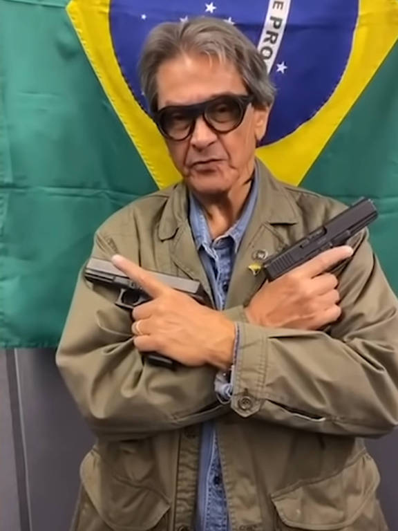 Um homem de óculos escuros está posando com duas armas cruzadas em frente ao peito. Ele está vestindo uma jaqueta verde e uma camisa jeans. Ao fundo, há uma bandeira do Brasil.