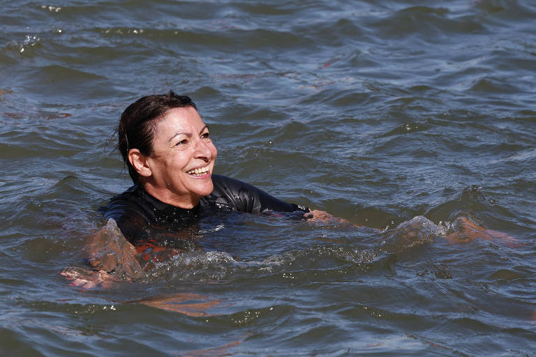 Uma mulher está nadando, com um sorriso no rosto. Ela veste uma roupa de mergulho preta e está parcialmente submersa na água. O mar ao seu redor está calmo, com pequenas ondulações.
