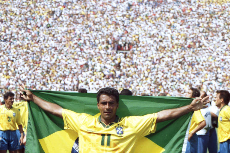 A imagem mostra um jogador de futebol brasileiro segurando a bandeira do Brasil aberta atrás de si. Ele está vestindo o uniforme da seleção brasileira, que consiste em uma camisa amarela com o número 11, shorts azuis e meias brancas. Ao fundo, há outros jogadores e uma grande multidão de torcedores nas arquibancadas.