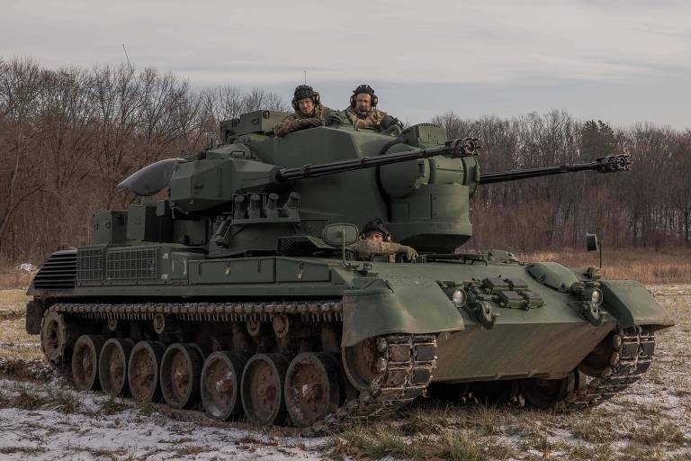 Em um campo nevado com árvores atrás está um veículo militar verde com dois canhões e três soldados.