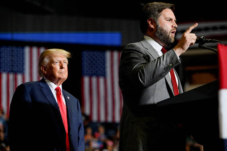 A imagem mostra dois homens em um comício. O homem à esquerda está de pé, vestindo um terno azul e gravata vermelha, enquanto o homem à direita está falando ao microfone, vestindo um terno cinza e gravata vermelha. Ao fundo, há duas bandeiras dos Estados Unidos penduradas.