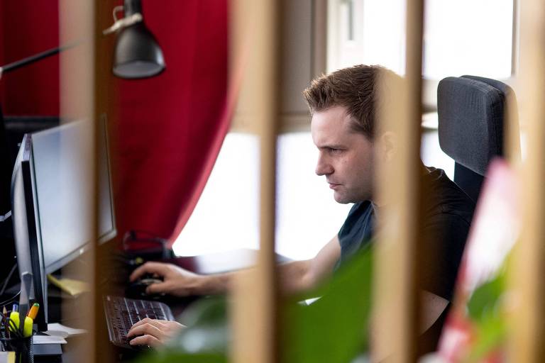 A imagem mostra um homem sentado em uma cadeira de escritório, trabalhando em um computador. Ele está concentrado na tela, com uma mão no teclado e a outra no mouse. Ao fundo, há uma cortina vermelha e uma luminária de mesa preta. A imagem é parcialmente obstruída por barras verticais desfocadas em primeiro plano.