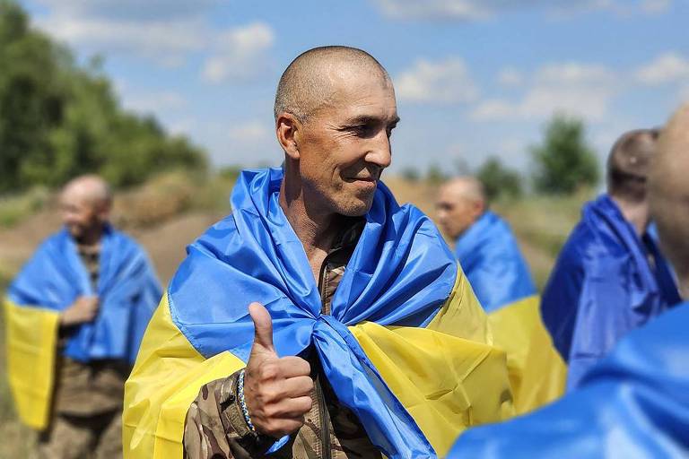 A imagem mostra um homem careca envolto em uma bandeira da Ucrânia, sorrindo e fazendo um gesto de positivo com a mão. Ao fundo, há outras pessoas também envoltas em bandeiras ucranianas, em um ambiente ao ar livre com céu azul e algumas árvores.
