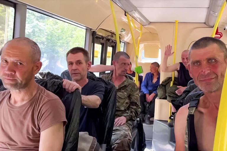 A imagem mostra um grupo de homens sentados dentro de um ônibus. Alguns estão vestindo roupas militares, enquanto outros estão com roupas casuais. O ambiente parece iluminado pela luz natural que entra pelas janelas do ônibus. Alguns dos homens estão olhando diretamente para a câmera, enquanto outros estão conversando entre si.