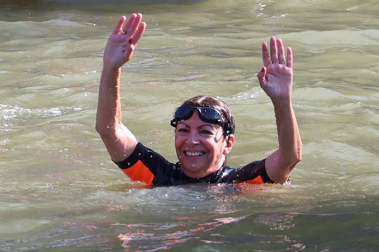 Uma pessoa está nadando em uma área de água, vestindo um traje de mergulho preto com detalhes laranja e óculos de natação. Ela está sorrindo e levantando ambas as mãos acima da água.
