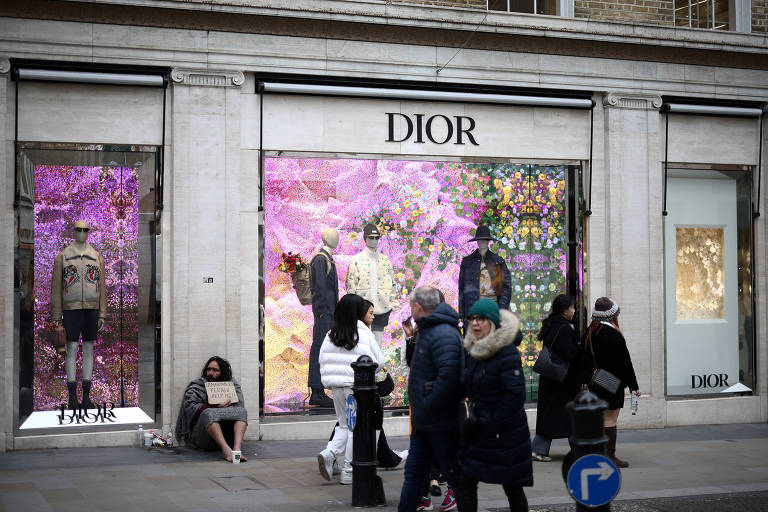 A imagem mostra a fachada de uma loja da Dior com grandes vitrines decoradas com fundos florais coloridos. Há manequins exibindo roupas da marca nas vitrines. Algumas pessoas estão caminhando na calçada em frente à loja, e uma pessoa está sentada no chão ao lado da vitrine à esquerda.