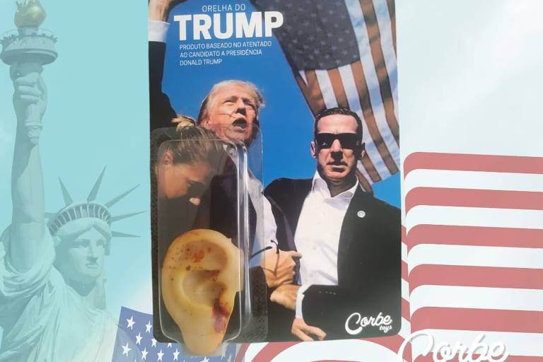 A imagem mostra um produto chamado 'Orelha do Trump'. A embalagem apresenta uma foto de duas pessoas, uma delas levantando um dos braços com o punho cerrado e a outra usando óculos escuros, com uma bandeira dos Estados Unidos ao fundo. Na parte inferior esquerda da embalagem, há uma orelha de plástico. No fundo da imagem, há uma estátua da liberdade e uma bandeira dos Estados Unidos estilizada. O texto na parte superior da imagem diz 'ORELHA DO TRUMP'.