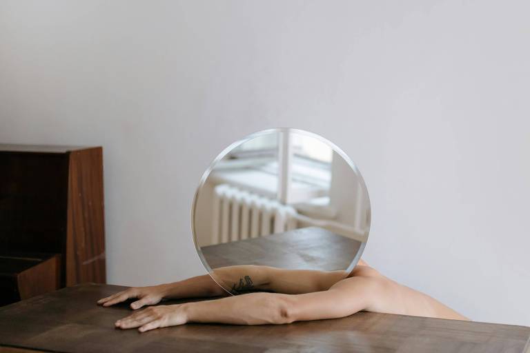 pessoa deitada sobre mesa de madeira; sobre seu rosto há o um espelho redondo
