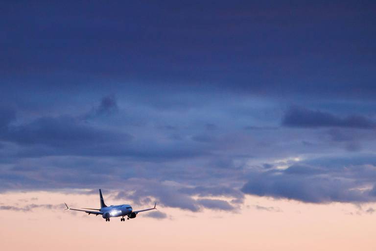 A imagem mostra um avião em voo, com as luzes acesas, contra um céu com nuvens escuras e um horizonte em tons de rosa e azul 
