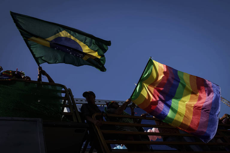A imagem mostra duas bandeiras sendo erguidas contra um céu azul. À esquerda, há uma bandeira do Brasil, com seu design verde, amarelo e azul. À direita, há uma bandeira do orgulho LGBTQIA+, com listras coloridas em vermelho, laranja, amarelo, verde, azul e roxo. As bandeiras estão sendo seguradas por pessoas que não são claramente visíveis.