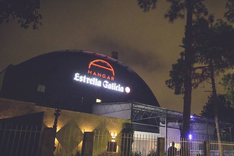 A imagem mostra a entrada de um edifício à noite, com uma cúpula iluminada e árvores ao redor. Há uma placa iluminada na cúpula com o texto 'Hangar'. A iluminação do edifício é amarela e há luzes verdes iluminando as árvores à direita.