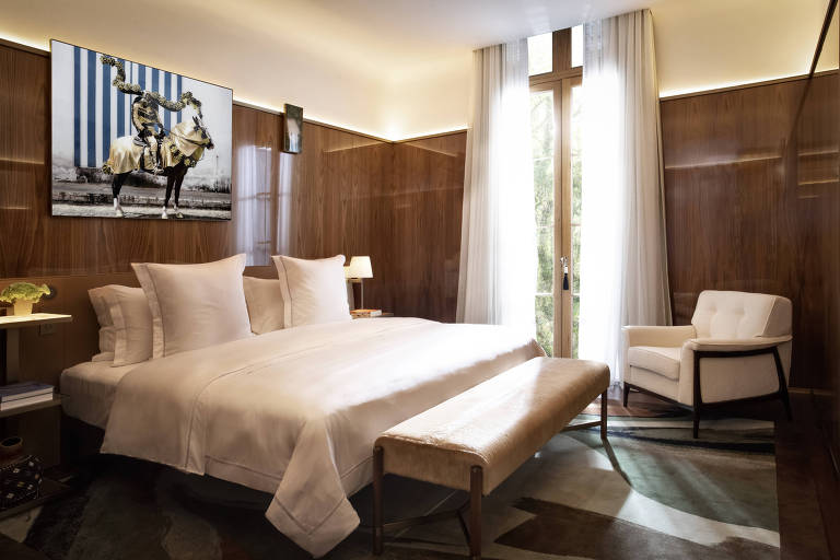 Quarto de hotel com cama king size e lençóis brancos, paredes de madeira, uma porta de varanda com cortina clara e um quadro na parede
