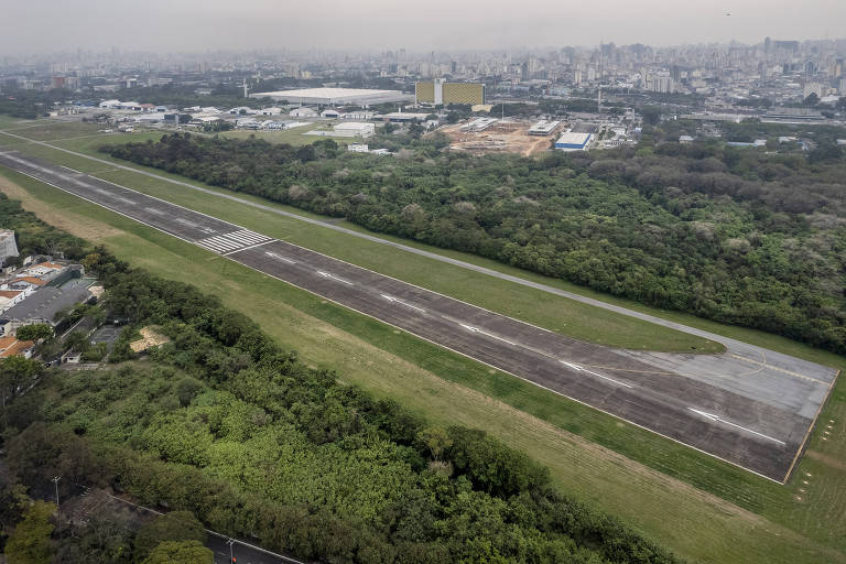 Imagem aérea mostra pista do aeroporto com densa vegetação nas margens e acidade ao fundo