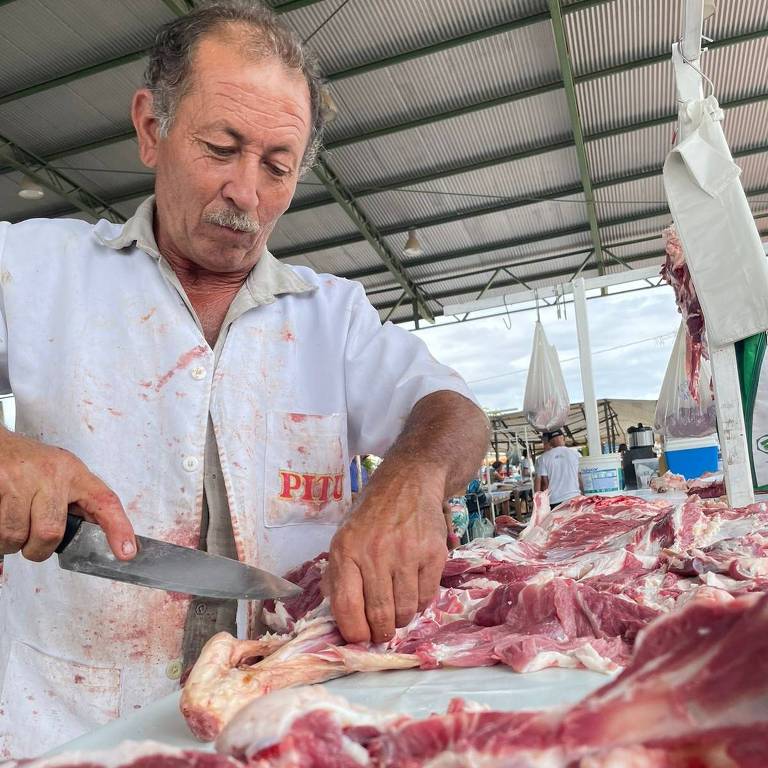 A imagem mostra um açougueiro usando uma faca grande para cortar pedaços de carne em um mercado coberto. Ele está vestindo um jaleco branco e parece concentrado em seu trabalho. Há várias peças de carne crua espalhadas sobre a mesa à sua frente.