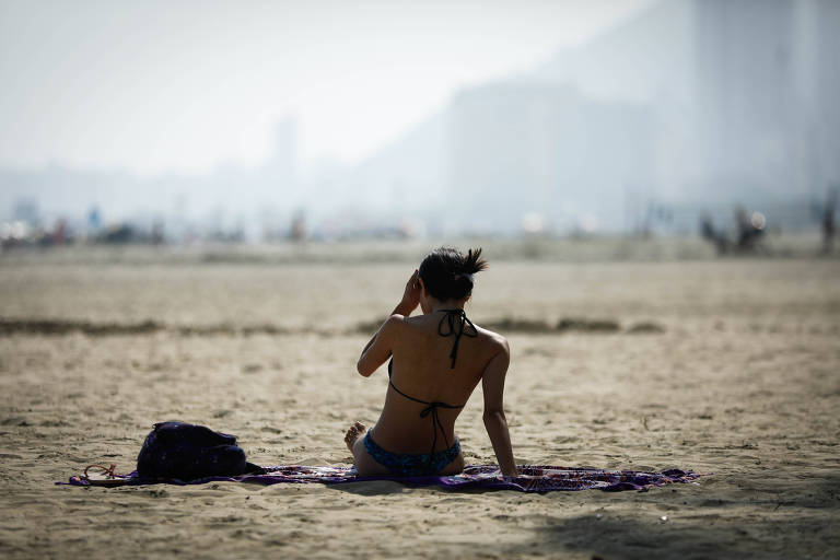 A imagem mostra uma pessoa sentada na areia de uma praia, de costas para a câmera. Ela está usando um biquíni e parece estar mexendo no cabelo. Ao lado dela, há uma bolsa preta. Ao fundo, a praia está levemente desfocada, com algumas pessoas e construções visíveis à distância