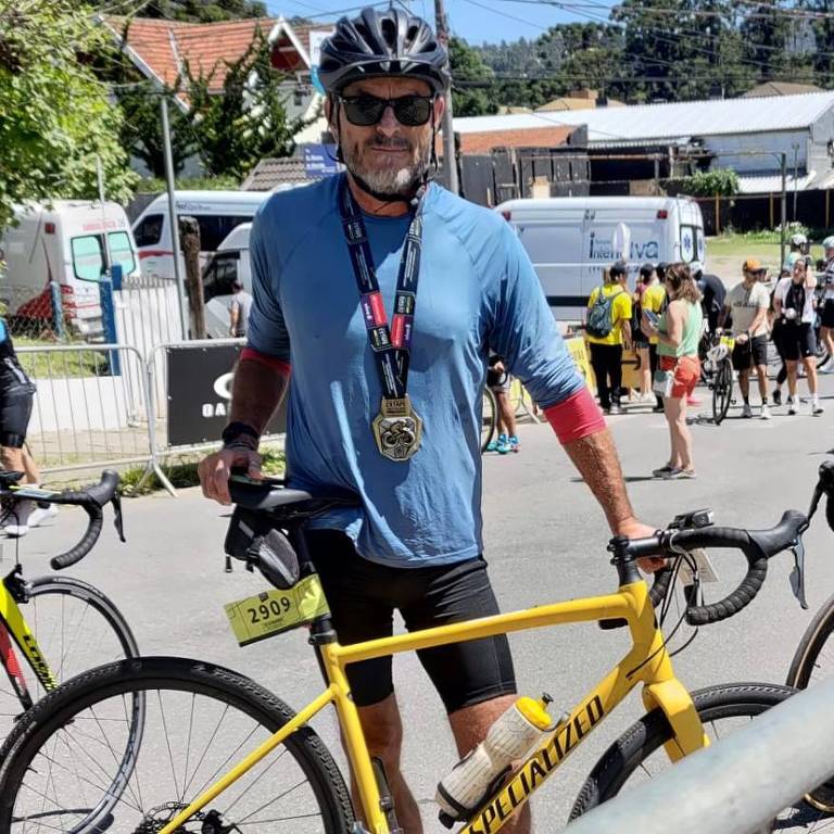 Um ciclista está posando ao lado de uma bicicleta amarela em um ambiente ao ar livre. Ele está usando um capacete preto, óculos de sol, uma camiseta azul e shorts pretos. Ao fundo, há outras pessoas, bicicletas e algumas construções, incluindo uma tenda branca.