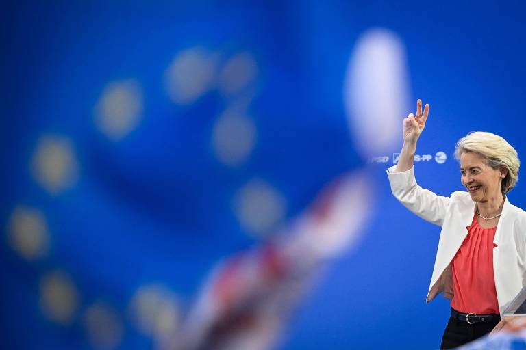 A imagem mostra Ursula von der Leyen, uma pessoa loira acenando com a mão direita, vestindo um blazer branco e uma blusa vermelha. Ao fundo, há uma bandeira da União Europeia desfocada, com um fundo azul e estrelas amarelas dispostas em círculo.