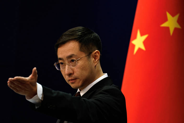 Um homem de óculos e terno preto está gesticulando com a mão direita estendida para frente. Atrás dele, há uma bandeira da China com fundo vermelho e estrelas amarelas.