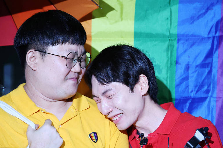 A imagem mostra duas pessoas abraçadas em frente a uma bandeira do arco-íris. A pessoa à esquerda usa uma camisa amarela e óculos redondos, enquanto a pessoa à direita usa uma camisa vermelha e parece estar emocionada, com os olhos fechados e um sorriso no rosto.