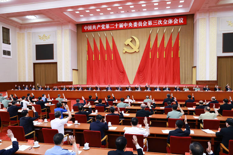 A imagem mostra uma reunião de autoridades em um grande salão. Há várias pessoas sentadas em mesas dispostas em formato de U, com algumas levantando as mãos. Ao fundo, há várias bandeiras vermelhas e um grande emblema dourado do martelo e foice, símbolo do Partido Comunista Chinês. Acima do emblema, há um banner vermelho com texto em chinês.
